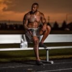 best sports supplements for recovery after football – Muscular athlete sitting on a bench with supplements, enjoying a sunset at an outdoor stadium.