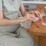prenatal vitamins Amazon Pakistan – Unrecognizable pregnant female sitting at wooden table with glass of water while taking prenatal vitamins in light kitchen at home