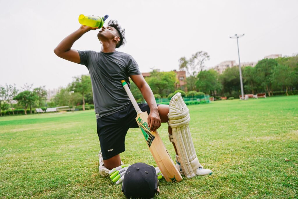 hydration supplements for cricket players Pakistan – Full length of ethnic male cricket player in sportswear with bat and helmet and pads kneeling on grass while drinking water from sport bottle