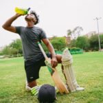 hydration supplements for cricket players Pakistan – Full length of ethnic male cricket player in sportswear with bat and helmet and pads kneeling on grass while drinking water from sport bottle