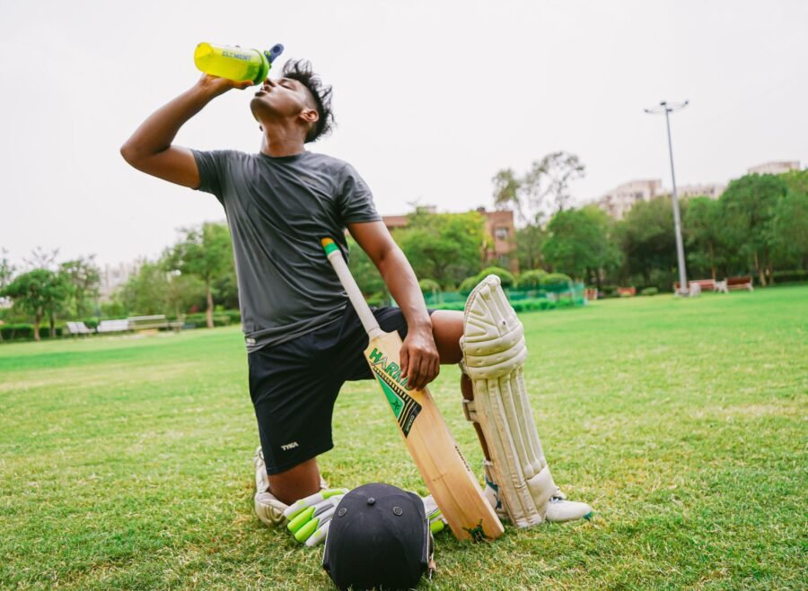 hydration supplements for cricket players Pakistan – Full length of ethnic male cricket player in sportswear with bat and helmet and pads kneeling on grass while drinking water from sport bottle