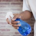stud spray price in pakistan – A close-up shot of a hand holding a blue spray bottle against a brick wall background.