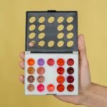 lip palette in pakistan – Hand holding a vibrant makeup palette with lipstick shades against a yellow backdrop.