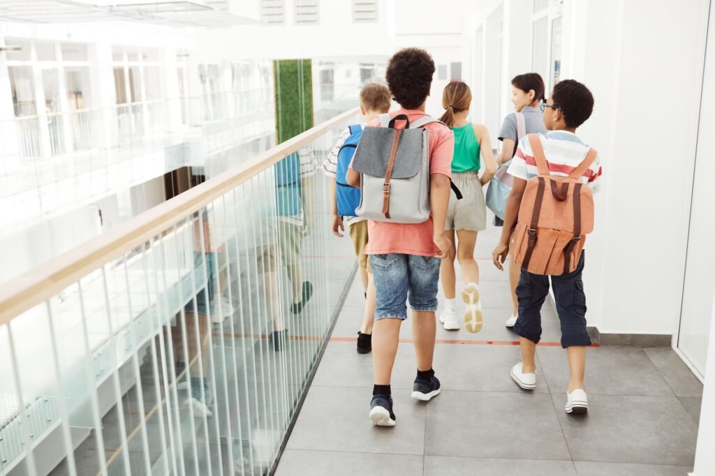 school bags with wheels – Group of children walking inside a modern school hallway with backpacks.
