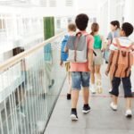 school bags with wheels – Group of children walking inside a modern school hallway with backpacks.