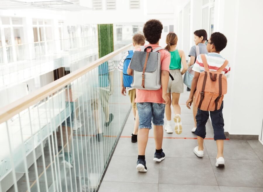 school bags with wheels – Group of children walking inside a modern school hallway with backpacks.